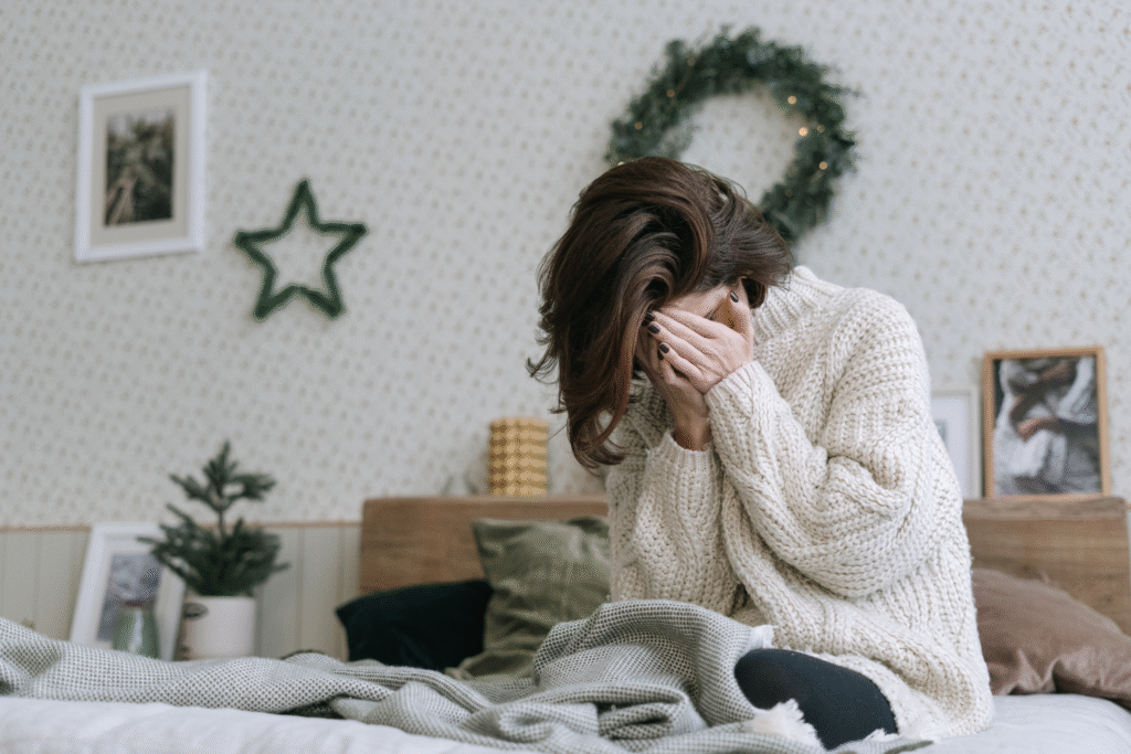 Women with head in hand looking upset and overwhelmed whilst sitting on bed. There are some Christmas decorations in the background.